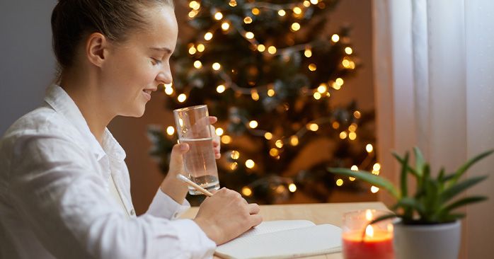 Woman journaling by the Christmas tree and sipping a glass of water.
