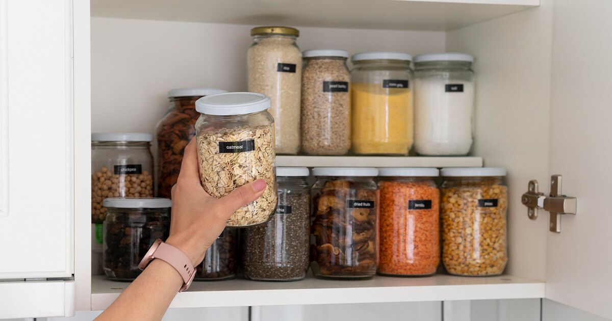 Woman pulling jar of oatmeal out of cupboard. Shelves of labelled pantry items in background.