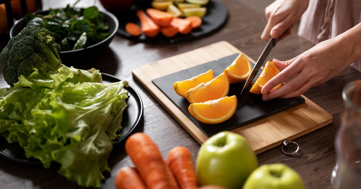 Woman cutting oranges on a cutting board. Variety of fruits and vegetables surrounding it.