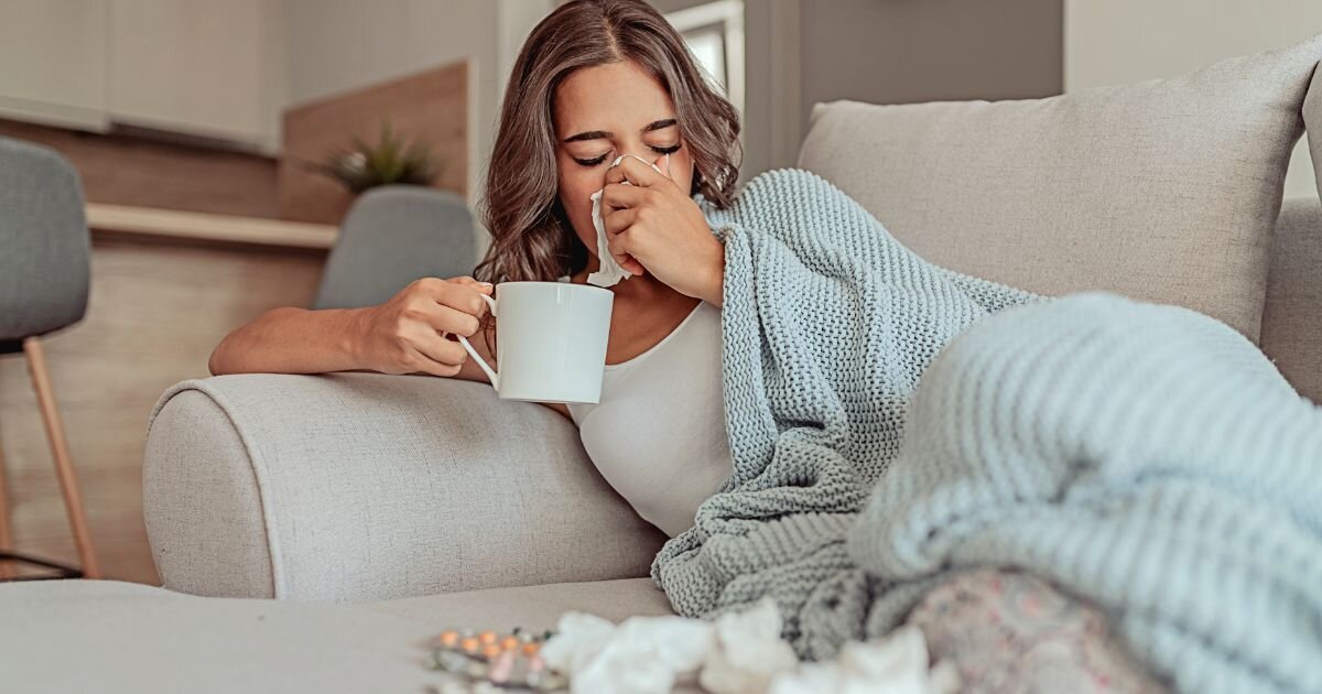 women laying on couch sick and blowing nose holding tea cup 